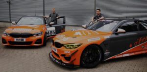 Zwei orangefarbene BMW-Rennwagen von Sonic Equipment mit Logo-Lackierung, stehen vor einer Industriehalle; Fahrer positionieren sich an den Fahrzeugen.