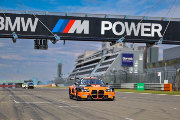 Ein orangefarbener BMW M4 Rennwagen fährt auf einer Rennstrecke unter einem BMW M Power Schild.