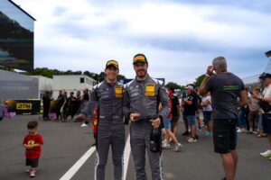 Zwei Rennfahrer in grauen DTM-Anzügen mit Helmen und Pokalen auf einer Rennstrecke.