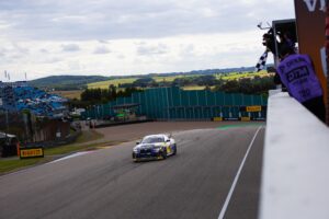 Ein Rennwagen fährt auf einer Rennstrecke während eines Motorsport-Events mit Zuschauern im Hintergrund.