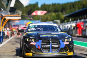 Ein blauer und schwarzer BMW M4 GT3 Rennwagen steht auf der Boxengasse. Die Scheinwerfer sind an, und man sieht das Ravenol-Logo auf der Motorhaube. Im Hintergrund sind undeutlich Personen und die Tribüne der Rennstrecke zu erkennen.