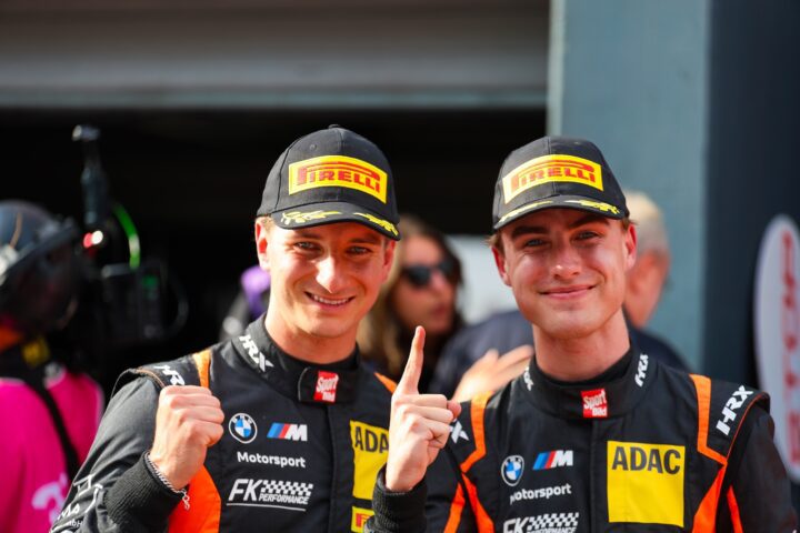 Zwei Rennfahrer in Pirelli-Caps und ADAC-Motorsportanzügen zeigen den Finger nach oben.