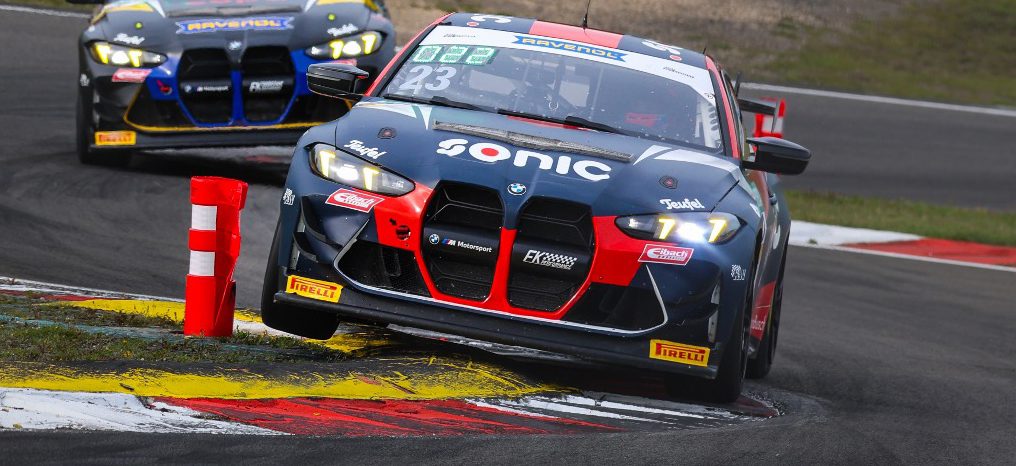 Zwei BMW M4 GT Rennwagen von FK Performance Motorsport, Nummer 23 in Dunkelblau/Rot und ein weiteres in Dunkelblau/Gelb, fahren dicht beieinander durch eine scharfe Kurve. Der vordere Wagen fährt über den rot-weiß-gelben Curb, was seine Aufhängung beansprucht.