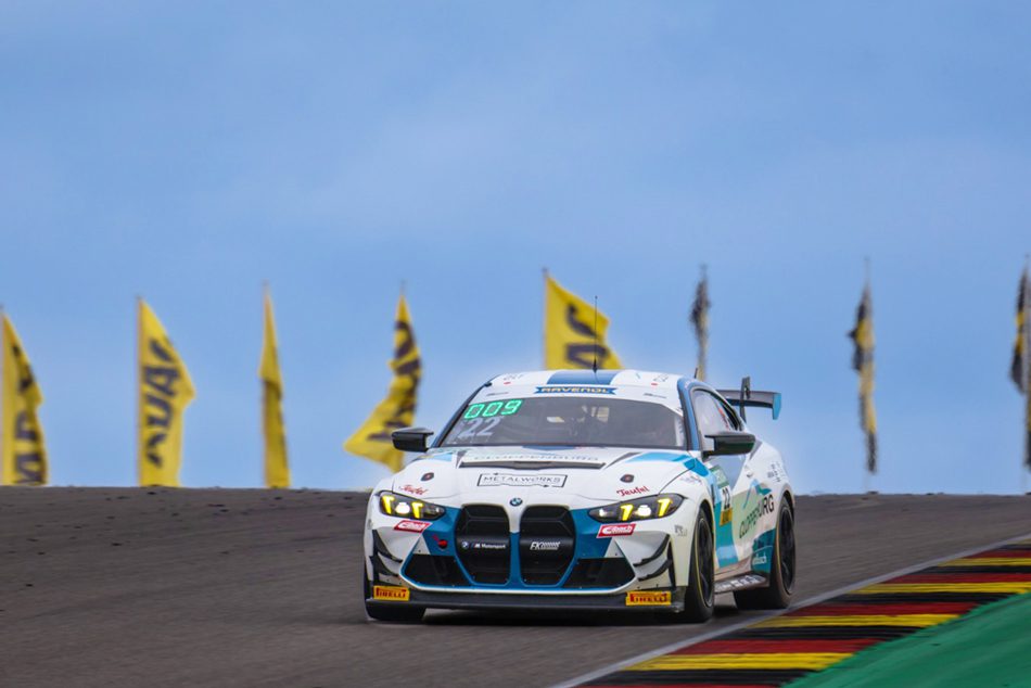 Ein weißer BMW M4 GT4 Rennwagen mit der Startnummer 009 des Teams FK Performance Motorsport, fährt eine leichte Steigung auf einer Rennstrecke. Im Hintergrund sind mehrere gelbe Sponsorenflaggen unter einem blauen, bewölkten Himmel zu sehen.