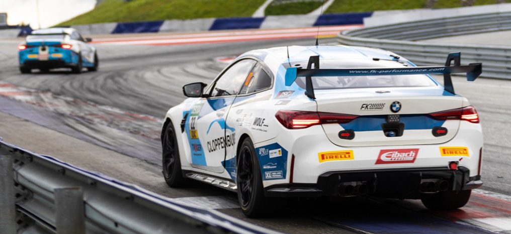 Ansicht des Hecks eines weißen BMW M4 GT4 EVO Rennwagens mit blauen Details und der Nummer 22 vom Team FK Performance Motorsport, der eine Kurve auf einer Rennstrecke nimmt. Sponsorenlogos und die Adresse der Team-Webseite sind auf der Heckscheibe und dem Kofferraumdeckel sichtbar.