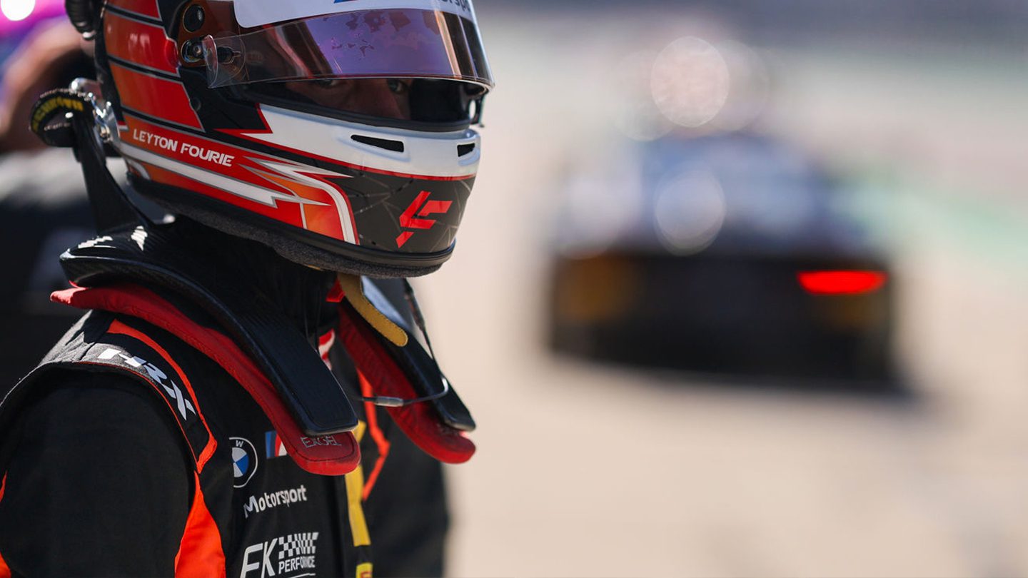 Nahaufnahme eines Rennfahrers, Leyton Fourie, in voller Ausrüstung (Helm mit Visier, HANS-System und Overall mit BMW Motorsport und FK Performance Logos), der in die Boxengasse blickt. Ein unscharfer Rennwagen ist im Hintergrund.