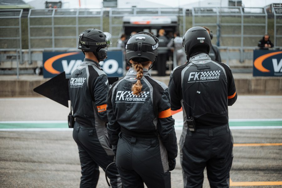 Drei FK Performance Crewmitglieder mit Helmen und Headsets stehen in der Boxengasse, einer hält ein Schild, Hintergrund mit Garage und VIVO-Werbung.