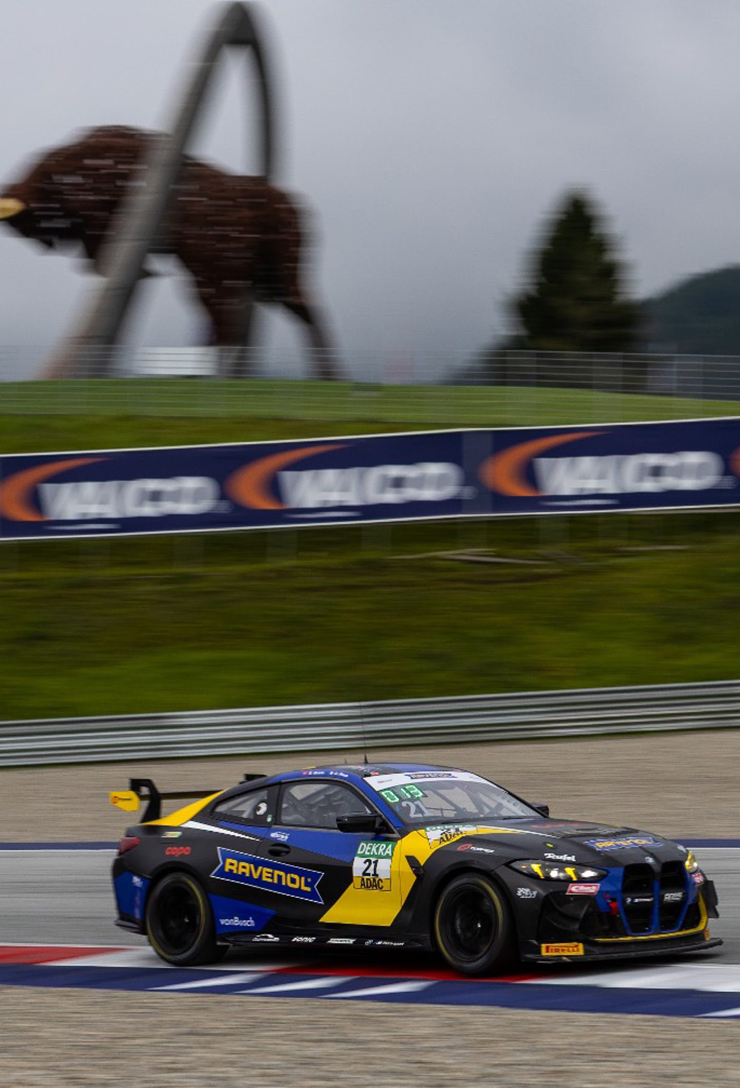 BMW M4 GT3 mit Startnummer 21 fährt auf dem Red Bull Ring, im Hintergrund die Stier-Skulptur, Sponsorenlogos und Streifenmarkierung am Streckenrand.