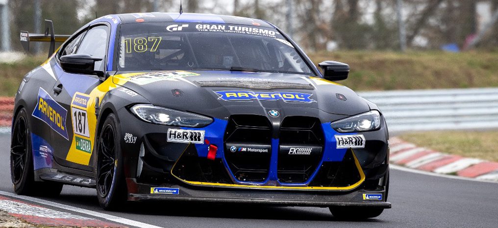Frontansicht eines schwarzen BMW M4 GT4 Rennwagens von FK Performance Motorsport mit gelben und blauen Akzenten, der die Startnummer 187 trägt und auf einer Rennstrecke fährt. Sponsorenlogos von Ravenol und Gran Turismo sind prominent auf der Motorhaube und der Windschutzscheibe zu sehen.