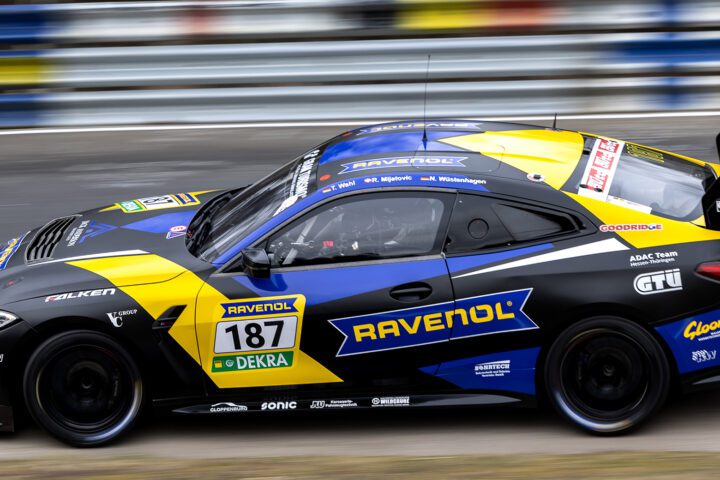 Dynamische Seitenansicht des BMW M4 GT4 Rennwagens (Startnummer 187, Ravenol/Falken) in schwarz-gelb-blauer Lackierung. Mitzieher-Aufnahme mit verschwommenem Hintergrund.