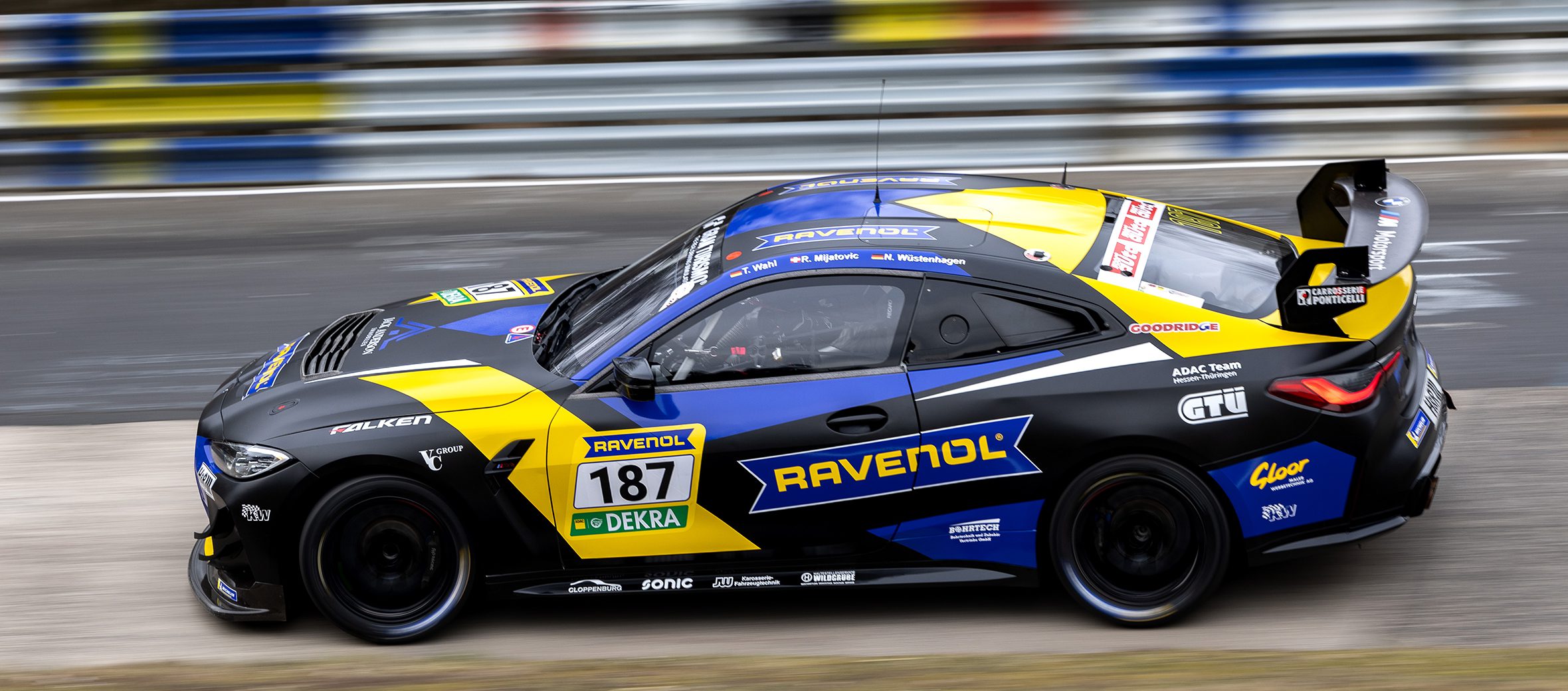 Dynamische Seitenansicht des BMW M4 GT4 Rennwagens (Startnummer 187, Ravenol/Falken) in schwarz-gelb-blauer Lackierung. Mitzieher-Aufnahme mit verschwommenem Hintergrund.