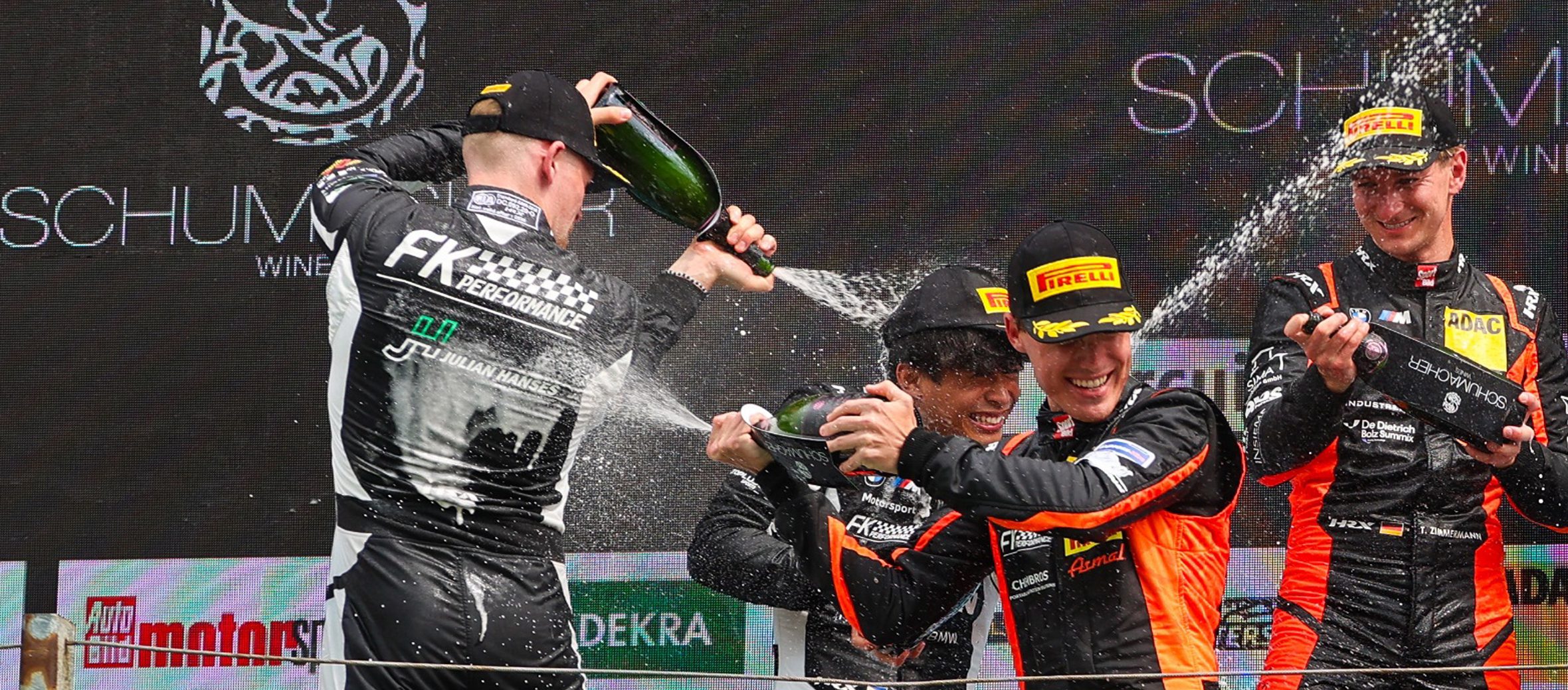 Vier Rennfahrer des Teams FK Performance auf dem Podium bei der Siegerehrung. Sie feiern mit einer ausgelassenen Sektdusche. Im Hintergrund Sponsoren-Logos.