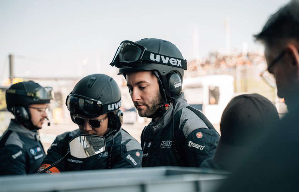 Nahaufnahme von drei Teammitgliedern von FK Performance Motorsport, die Helme mit uvex-Logos tragen und sich über ein Detail beugen. Die Szene spielt in der Boxengasse oder im Freien.