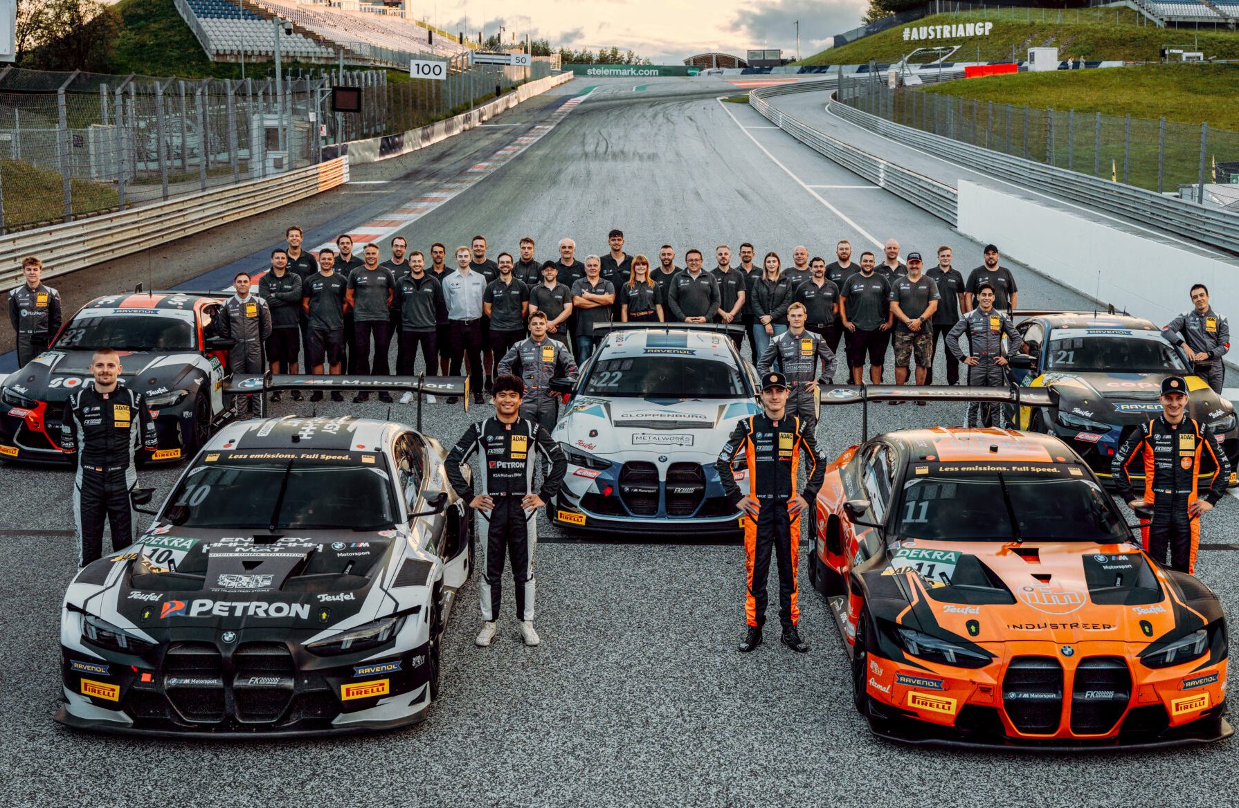 Gruppenfoto des FK Performance Motorsport Teams, inklusive zahlreicher Teammitglieder und mindestens vier BMW GT-Rennwagen (einige weiß/grau/schwarz, einer orange/schwarz, einer weiß/blau), aufgenommen auf einer Rennstrecke. Im Hintergrund ist die Beschriftung "AUSTRIAN GP" und eine "100"-Markierung der Strecke zu sehen.