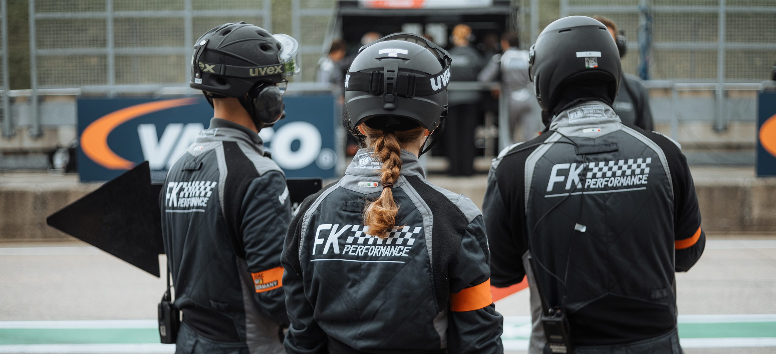 Rückenansicht von drei Teammitgliedern von FK PERFORMANCE, die in grauen Overalls mit dem Team-Logo und schwarzen Helmen in der Boxengasse stehen und auf die Strecke blicken. Die Overalls zeigen auch das Uvex-Logo auf den Helmen.