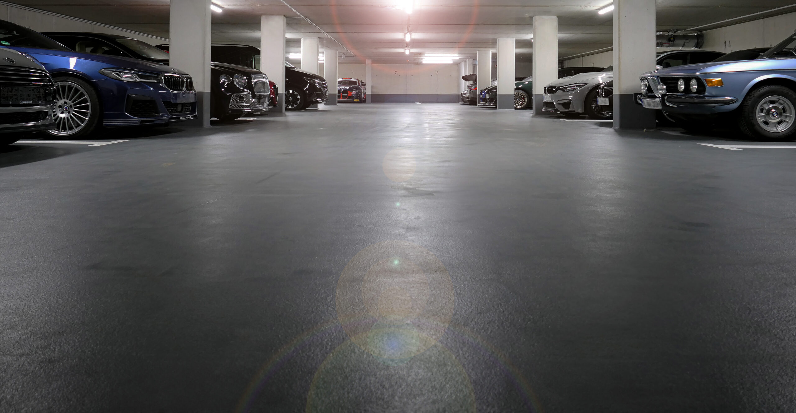 Innenansicht einer hell beleuchteten Tiefgarage mit Betonpfeilern. Verschiedene geparkte Autos, darunter moderne blaue BMWs und ein klassischer hellblauer Oldtimer.