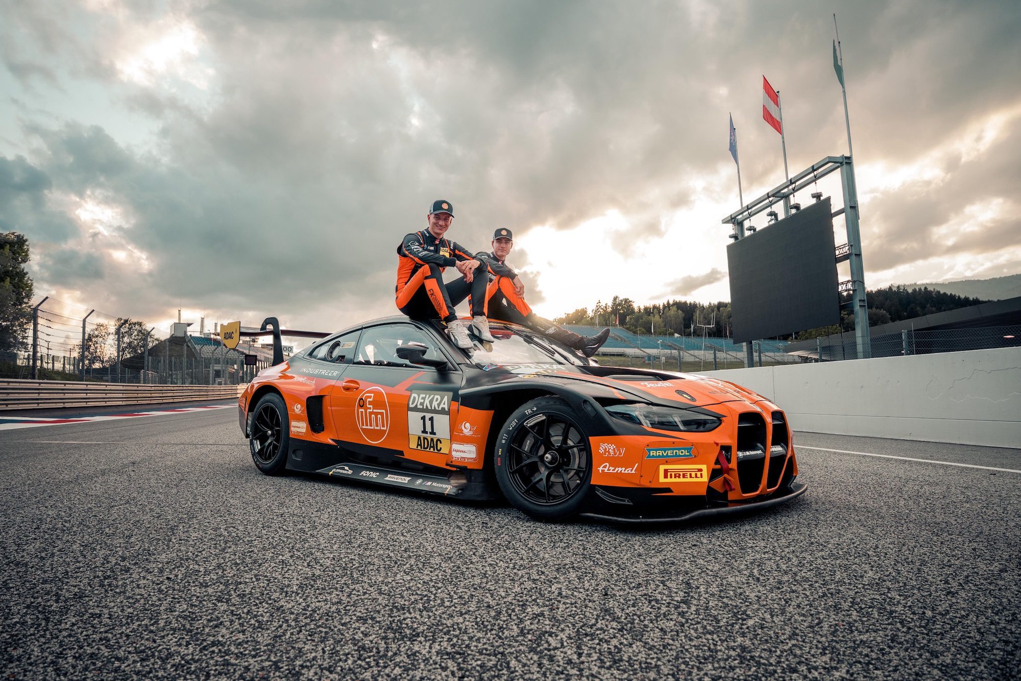 BMW M4 GT4 Rennwagen in Orange-Schwarz mit zwei Fahrern auf dem Dach auf einer Rennstrecke