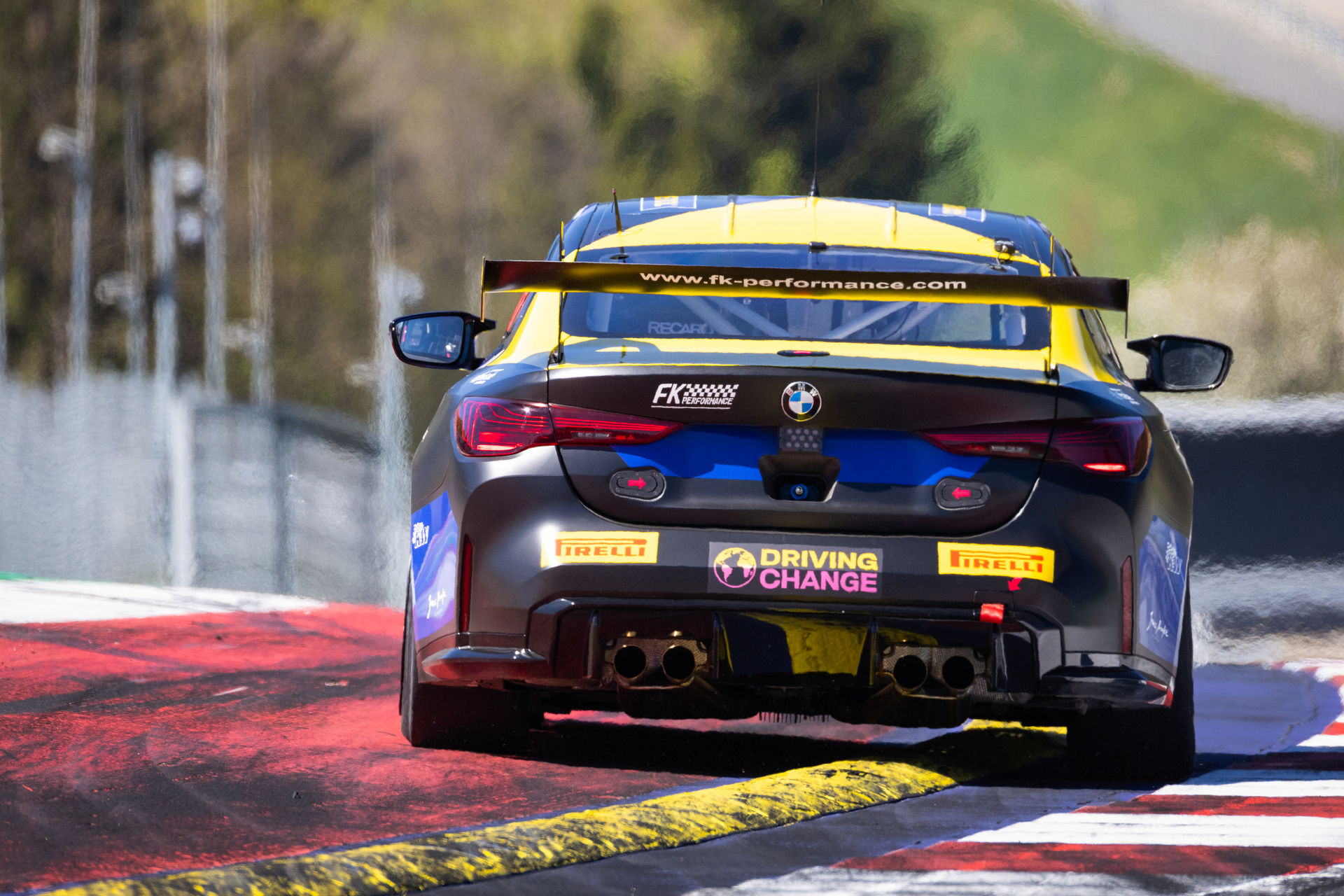 Heckansicht des schwarz-blau-gelben BMW M4 GT4 von FK Performance beim Überfahren der gelben Curbs am Kurvenausgang.