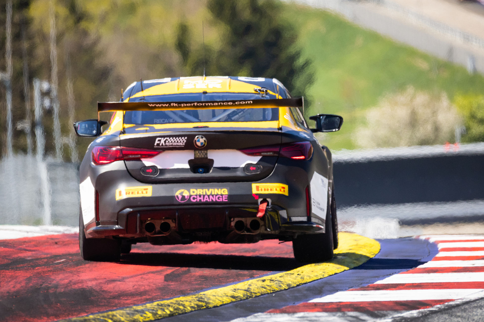 Heckansicht des schwarz-gelben BMW M4 GT4 von FK Performance Motorsport mit "Driving Change" Logo auf der Rennstrecke.