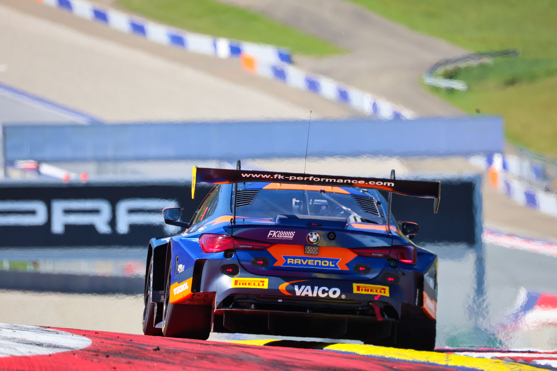 Heckansicht eines blau-orangenen BMW M4 GT3 Rennwagens von FK Performance mit massivem Heckflügel auf der Zielgeraden.