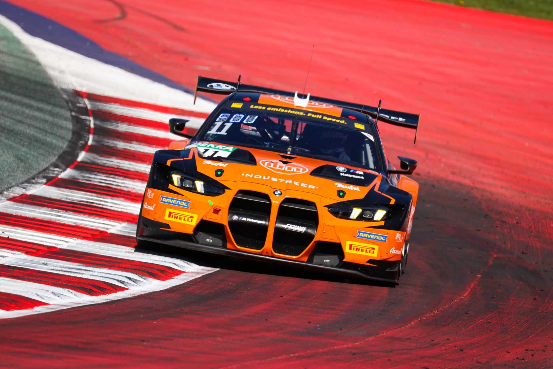 Frontalansicht des orangefarbenen BMW M4 GT3 (Startnummer 11) in einer schnellen Kurvenpassage auf rot-weißem Curb-Hintergrund.