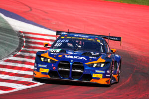 Frontansicht eines blau-orangenen BMW M4 GT3 Rennwagens von FK Performance (Nummer 10) in einer scharfen Linkskurve auf der Rennstrecke.