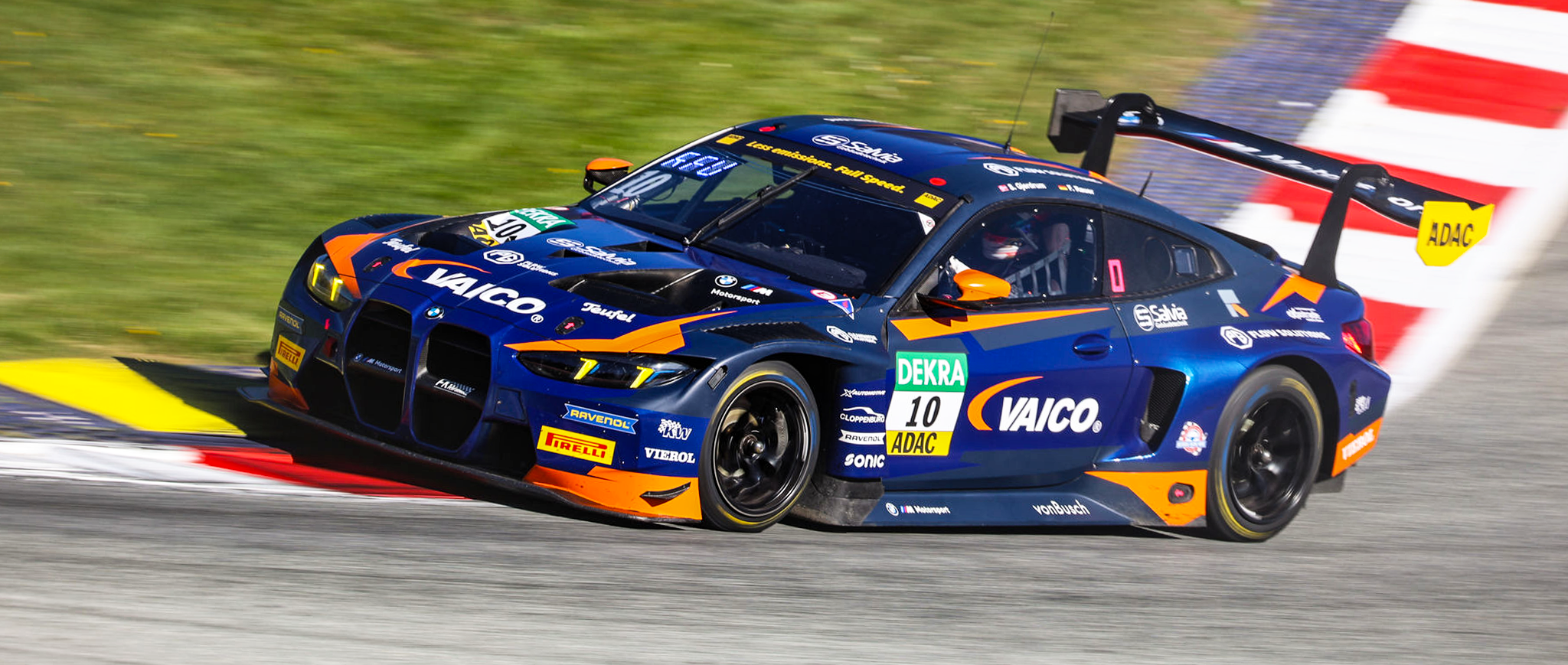 Seitenansicht des blau-orangenen BMW M4 GT3 Rennwagens von FK Performance (Nummer 10) beim Durchfahren einer Kurve auf der Rennstrecke.